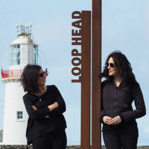 Two women posing at the Loop Head signage with the lighthouse in the background